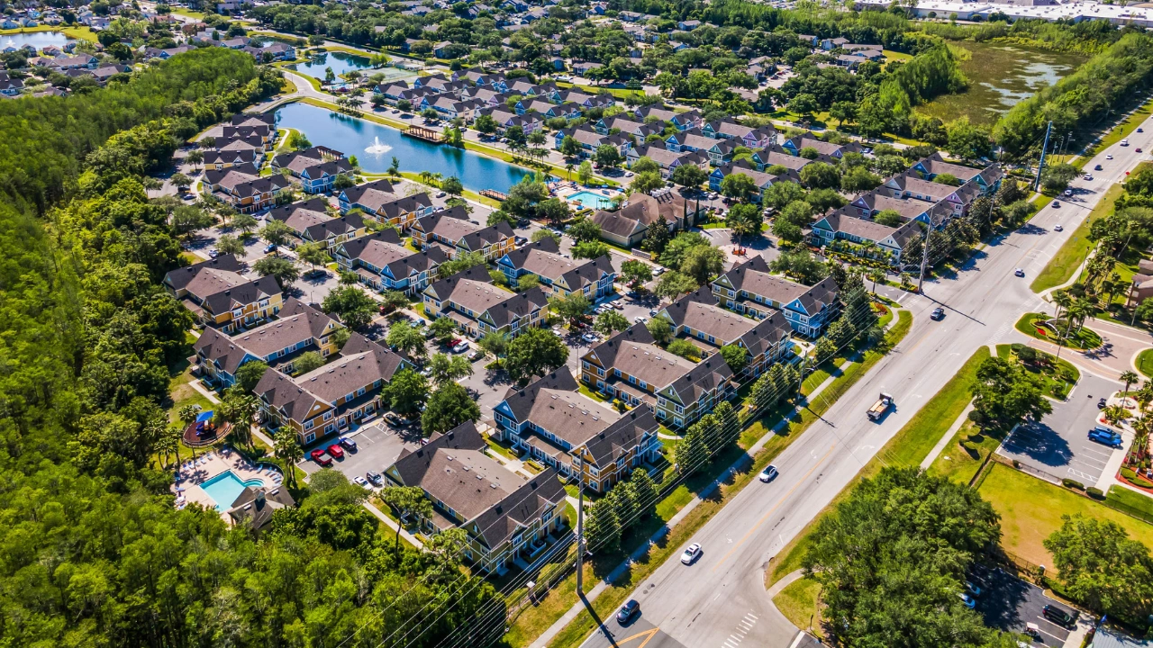 Aerial view of a beautifully designed residential community in Kissimmee, Florida, featuring colorful townhomes, lush green surroundings, a serene central pond, and well-maintained amenities, including a swimming pool and playground.