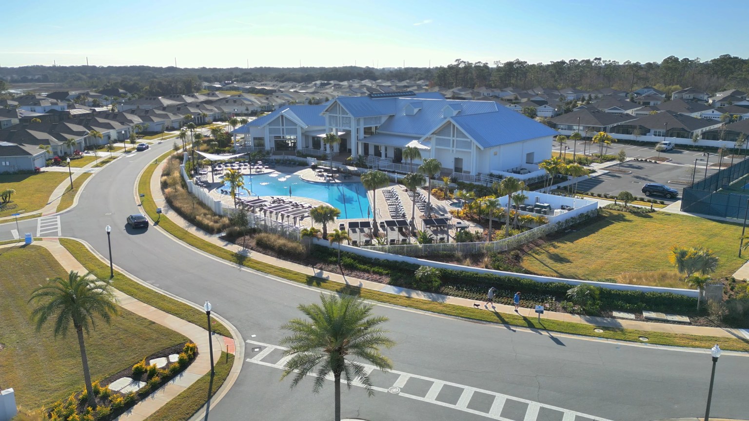 Aerial view of the Del Webb Oasis clubhouse with a blue metal roof, a resort-style pool with shade structures and loungers, courts at the right edge and surrounding homes, Winter Garden, Florida