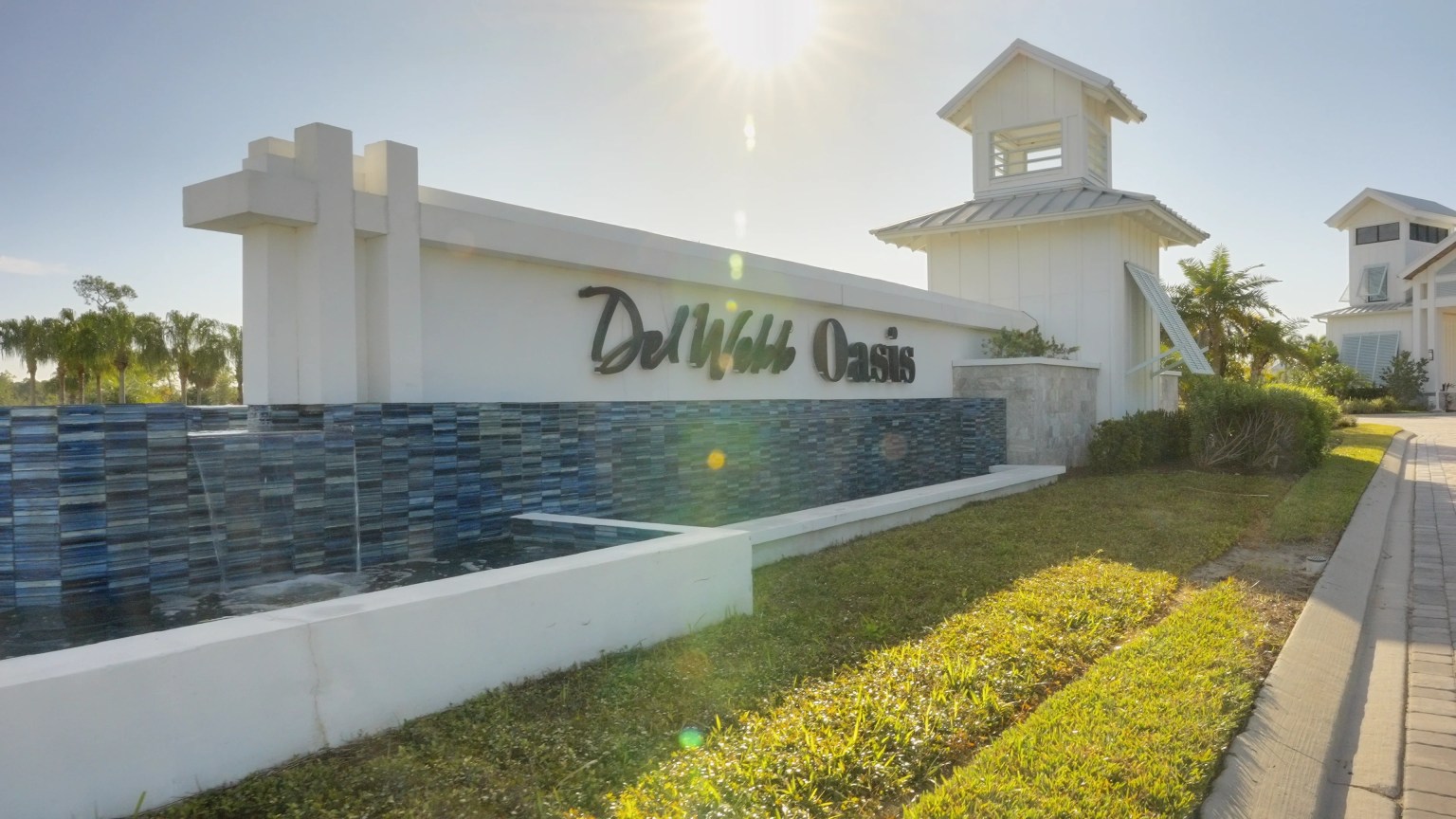 Del Webb Oasis entrance monument in Winter Garden, Florida: a white wall with the Del Webb Oasis script sign, a blue-tile water feature, a white cupola tower and palm trees under a sunny sky
