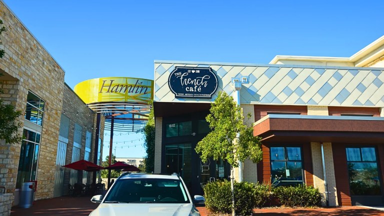The French Café storefront at Hamlin Town Center in Horizon West, Winter Garden, Florida, with its navy script sign, the yellow Hamlin tower sign behind it, a string-lit pergola, red patio umbrellas and stone and brick facades