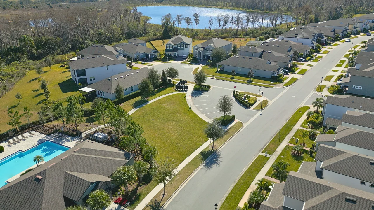 Aerial view of new construction single-family homes along a street with a community pool, open lawn and a lake behind them in Serenade at Ovation, Winter Garden FL