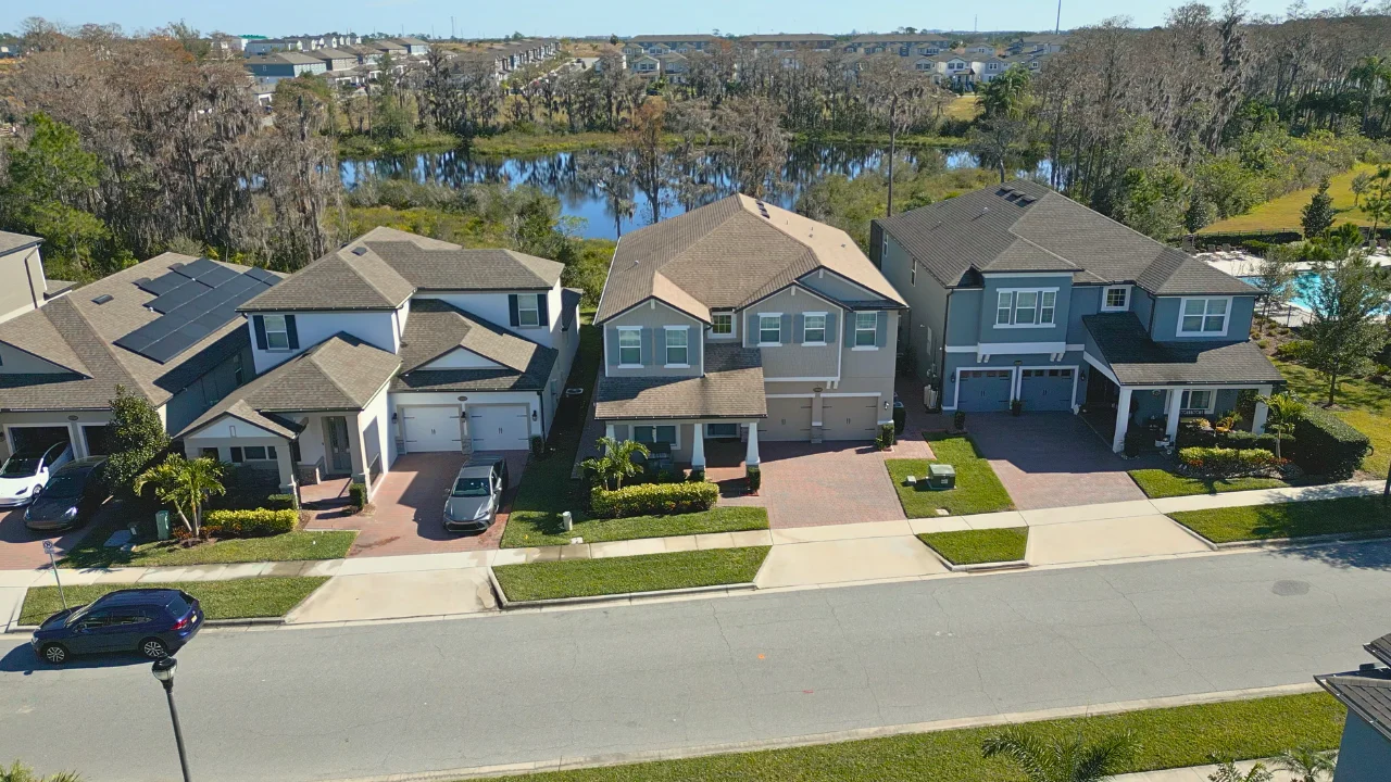 Aerial view of two-story single-family homes with paver driveways along a street in Serenade at Ovation, with a pond, trees and a community pool behind them, Winter Garden FL